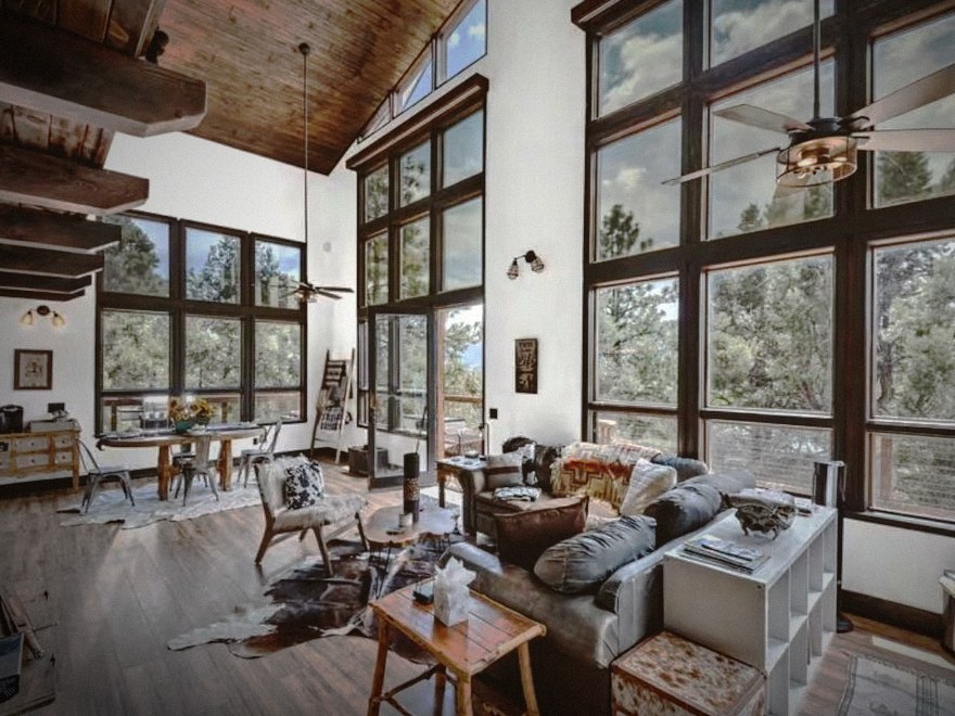 The great room of the original house: reclaimed timber beams, a vaulted wood ceiling, a hide rug on wide-plank floors, and a two-story wall of windows looking into ponderosa pines.
